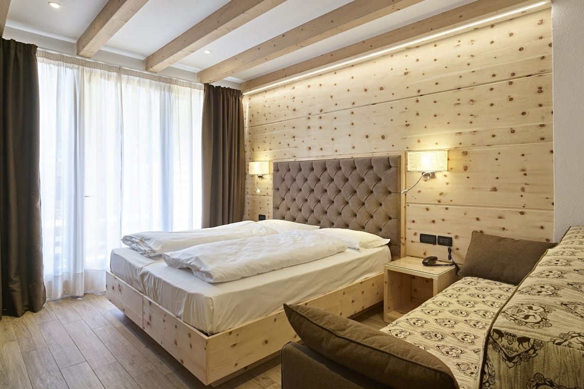 A hotel room with a large bed, a bedside table with a lamp, a wooden headboard, and a patterned sofa.