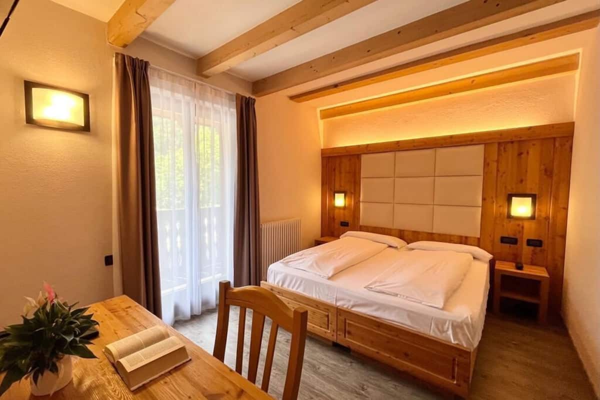 A bedroom with a wooden bed, bedside lamps, a window with curtains, and a wooden table with a plant.