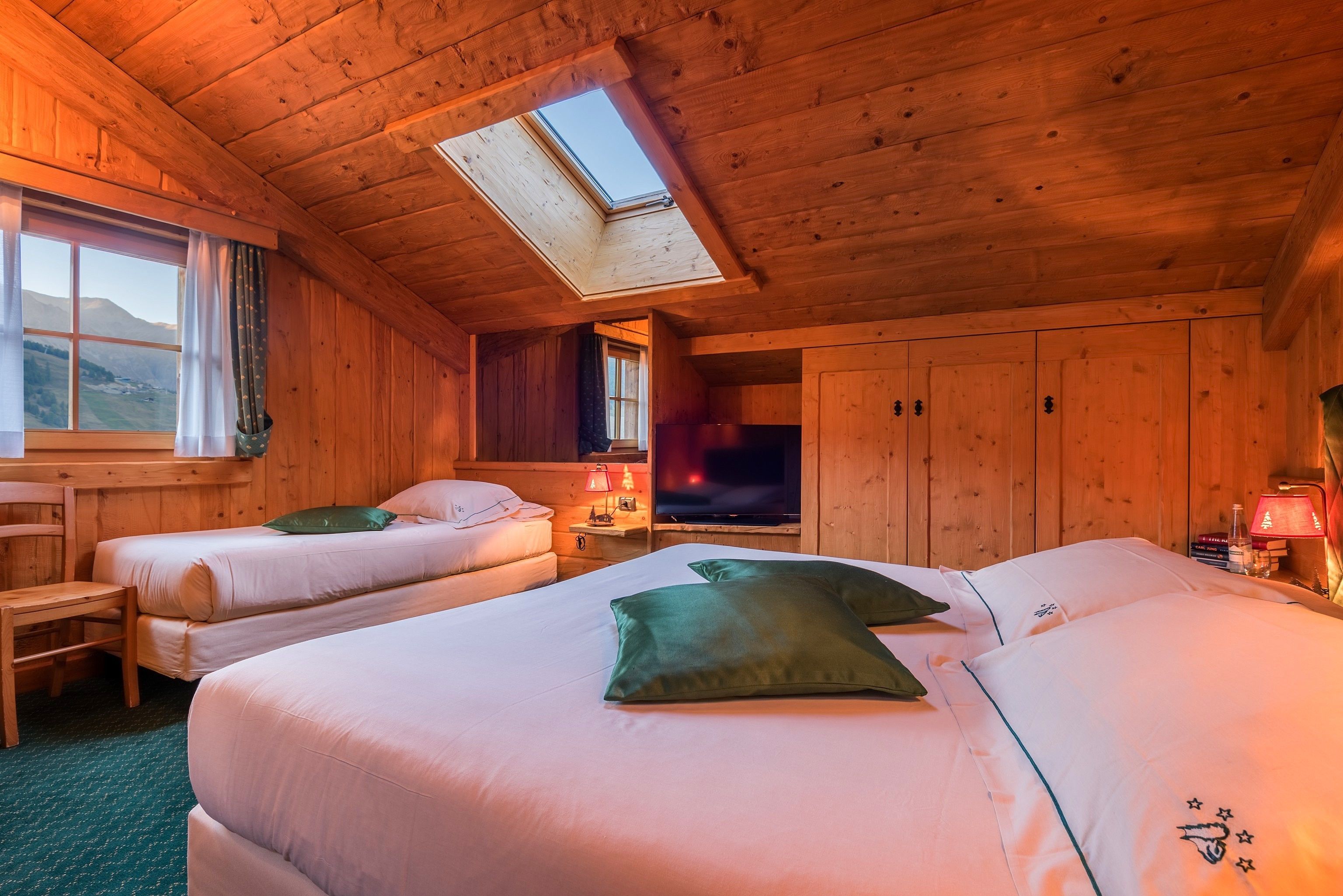 Ein gemütliches Holzblockhaus-Zimmer mit zwei Betten, einem Oberlicht, einem Fernseher und einem Fenster mit Blick ins Freie.
