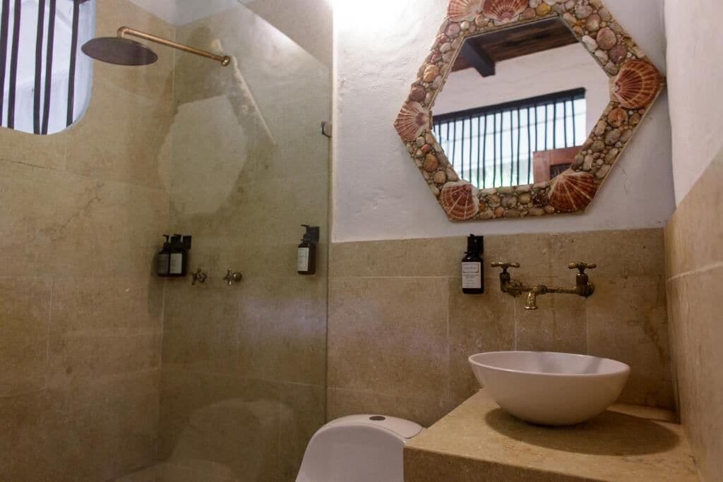 Un baño con lavamanos de piedra, ducha con puerta de cristal y espejo decorativo con conchas.