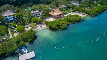 Vista aérea de un resort con playa, estructuras de techo de paja y un muelle con un bote.