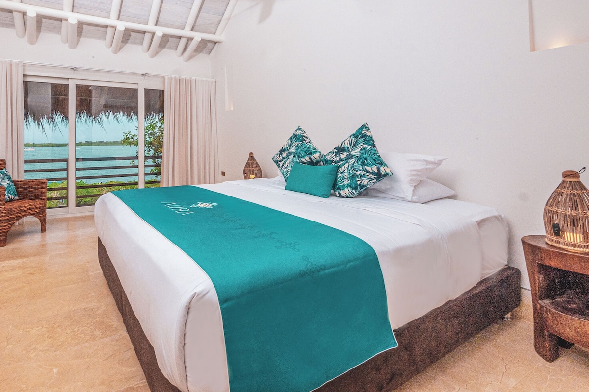 A bedroom with a large bed, a wicker chair, a nightstand with a lantern, and a view of the sea through a sliding door.