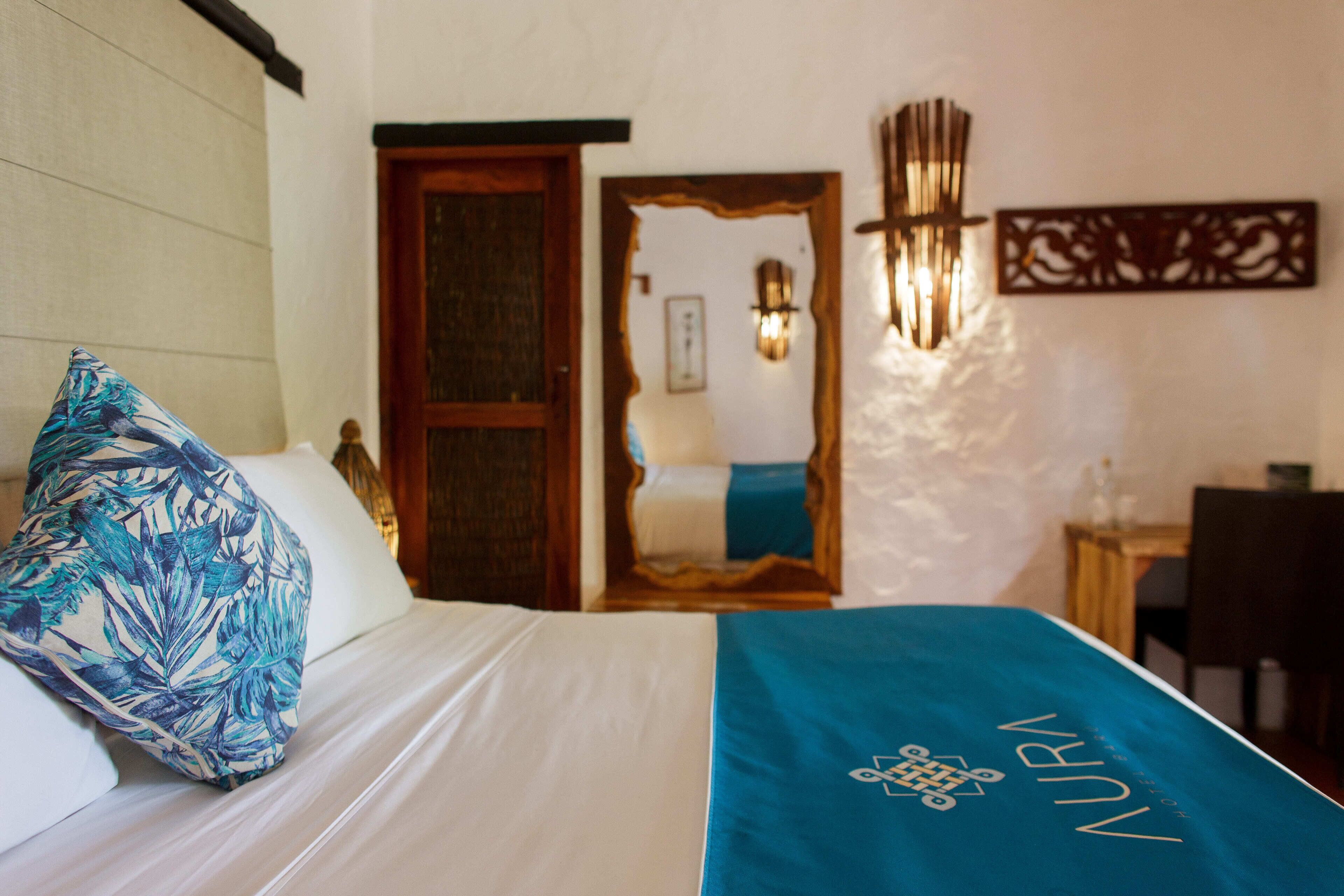Una habitación de hotel con una cama, una colcha azul con la palabra "NURN", una puerta de madera y una lámpara fijada a la pared.
