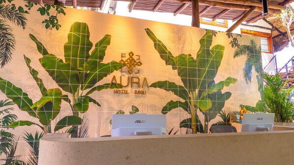 A reception area with a tropical theme, featuring large green leaves and the text "AURA HOTEL BARU".