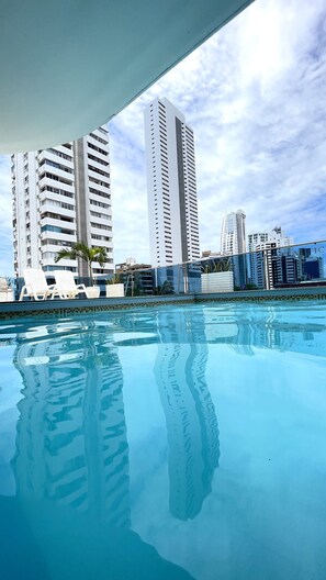 Piscina con agua cristalina que refleja edificios, rodeada de sillas de descanso y una palmera.