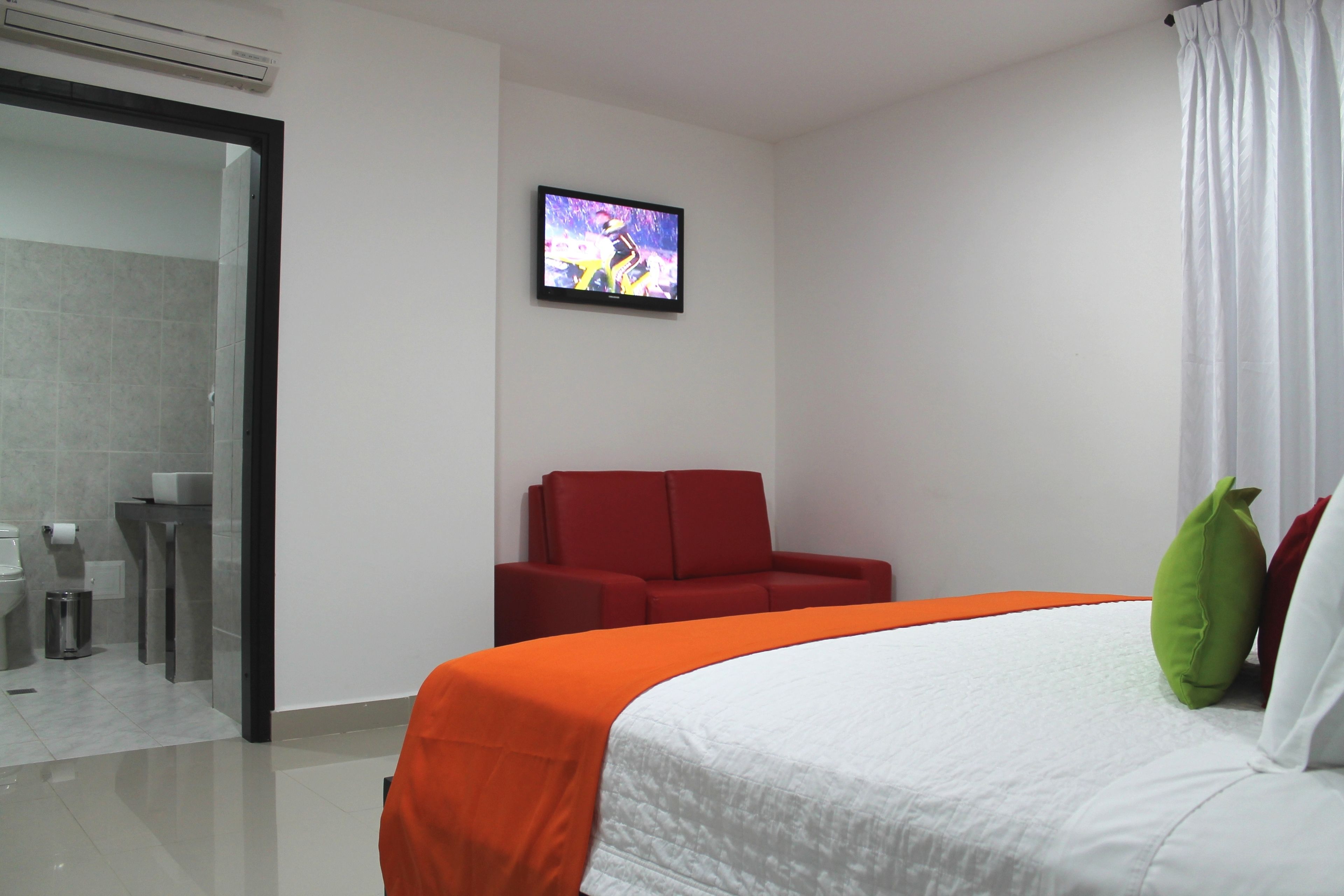 A hotel room with a bed, a TV, a red chair, and a bathroom visible through an open door.