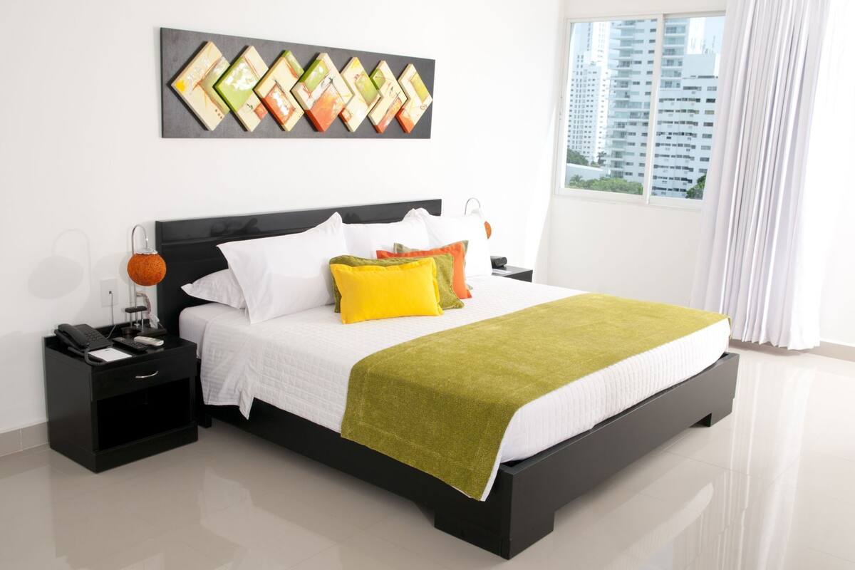A modern hotel room with a large bed, a nightstand, a lamp, and a view of the city.