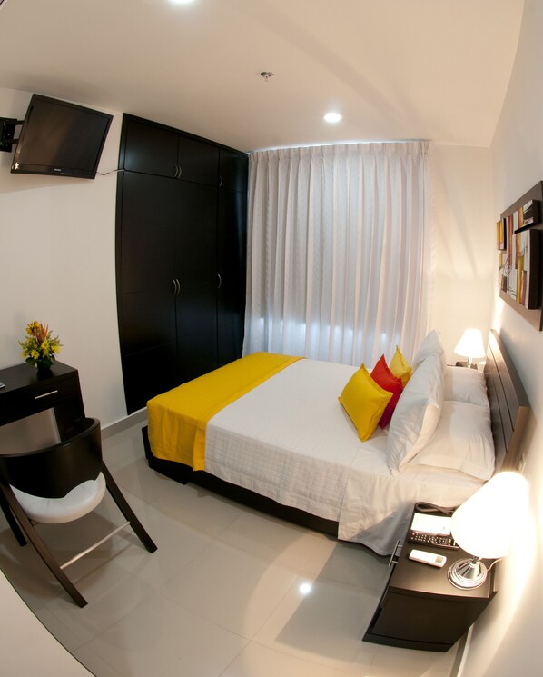 Una habitación de hotel moderna con una cama, un escritorio, una silla, un televisor montado en la pared y una mesita de noche con una lámpara.