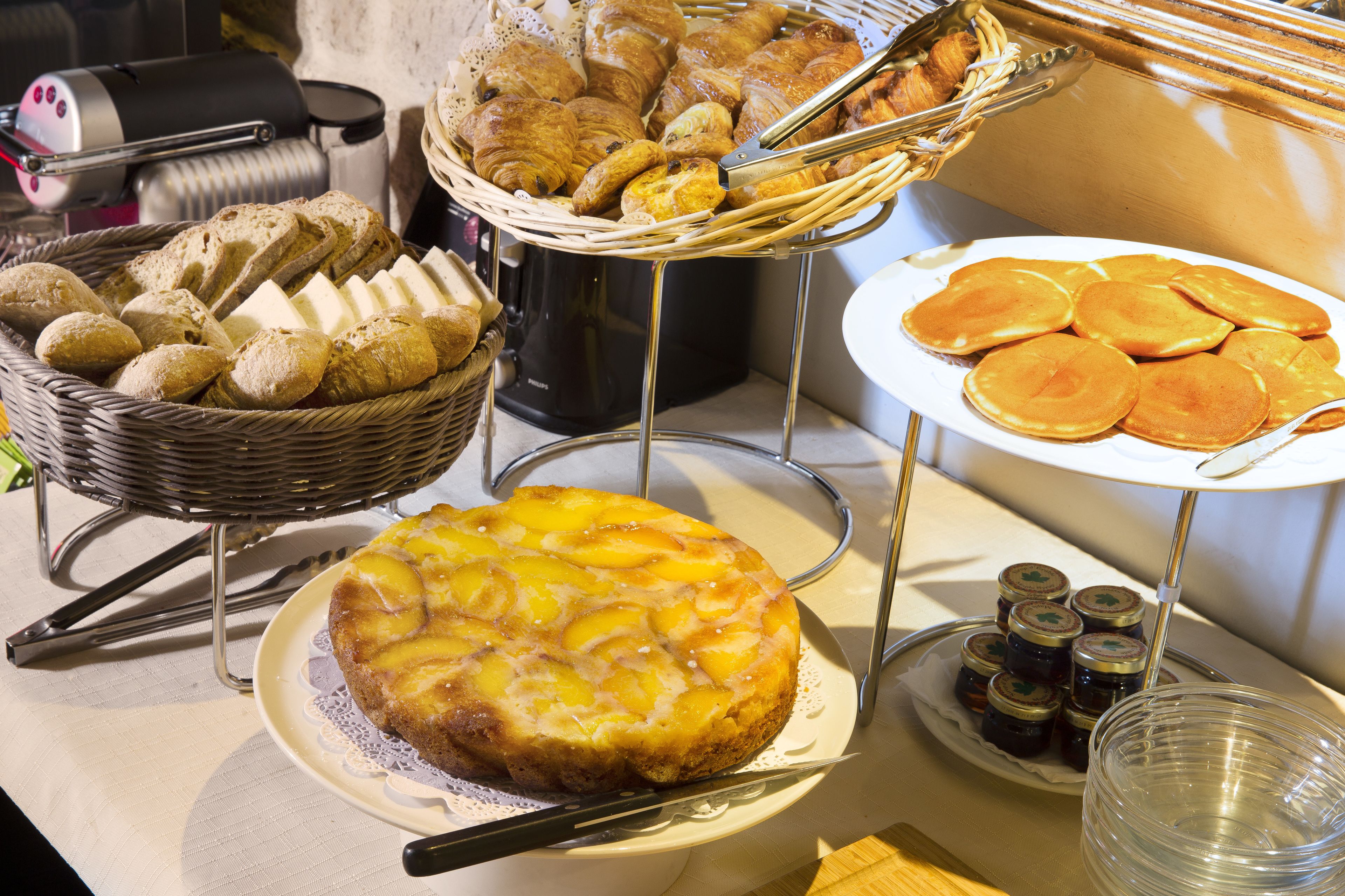A breakfast buffet with bread, pastries, pancakes, and a fruit-topped cake.