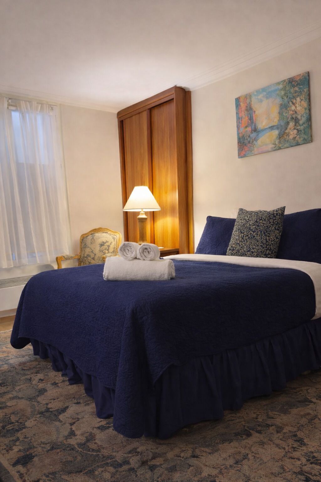 A hotel room with a large bed, a wooden wardrobe, a lamp, a chair, and a painting on the wall.