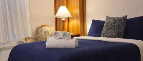 A hotel room with a large bed, a wooden wardrobe, a lamp, a chair, and a painting on the wall.