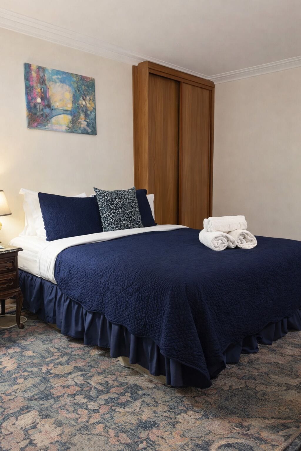 A neatly made bed with a dark blue bedspread and pillows, a wooden wardrobe, and a patterned carpet.