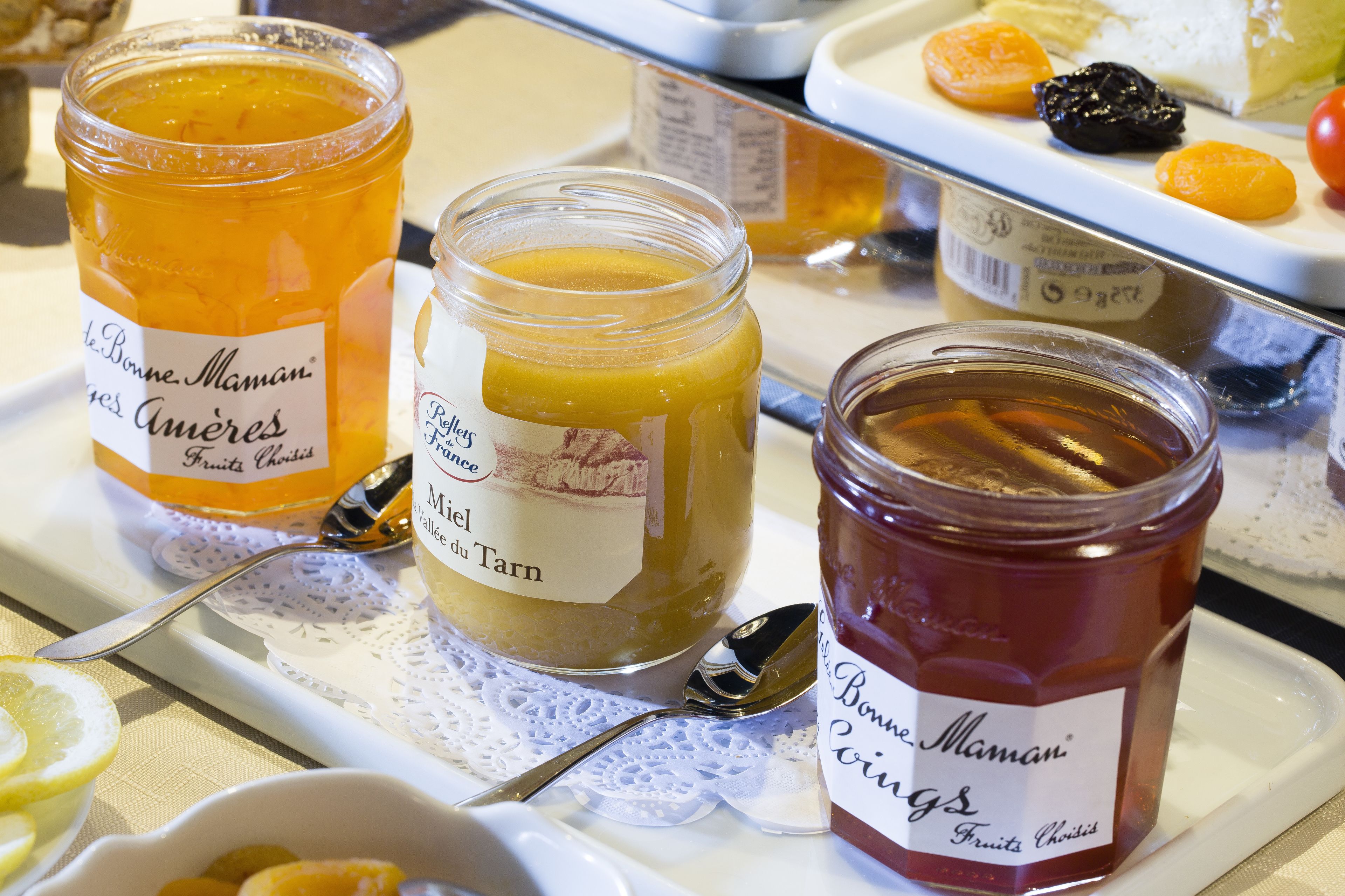 Three jars of fruit preserves on a tray with a spoon and a white bowl containing sliced lemons.