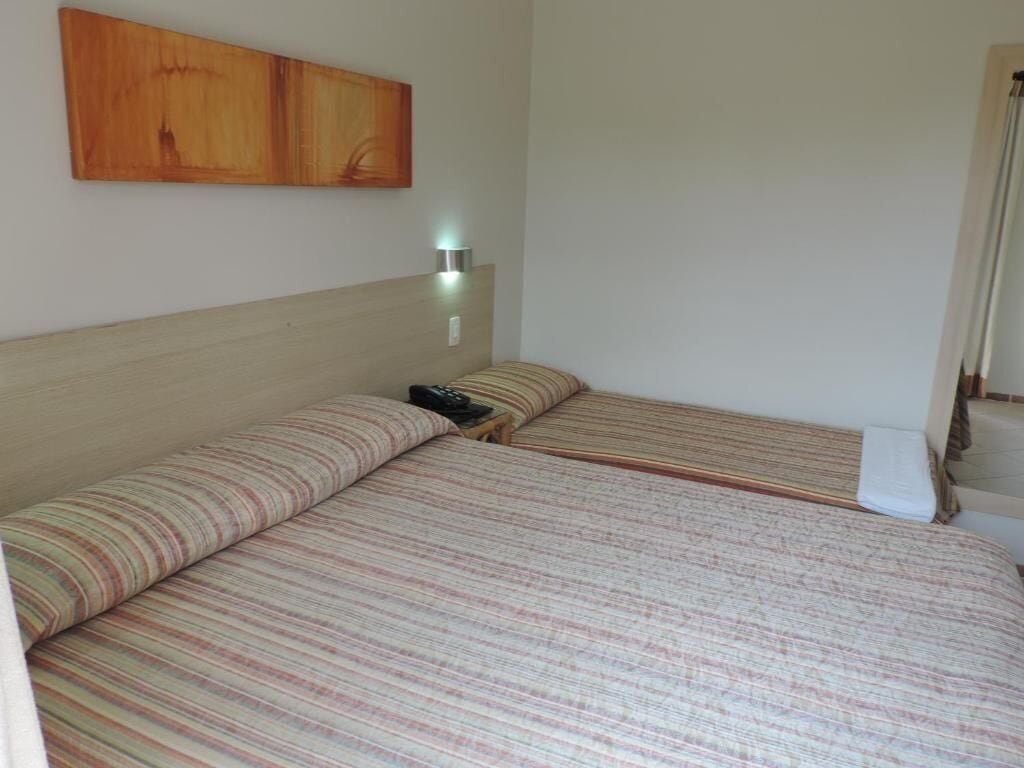 Quarto com duas camas, uma mesa de cabeceira, uma luminária na parede e uma porta.