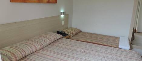 Quarto com duas camas, uma mesa de cabeceira, uma luminária na parede e uma porta.