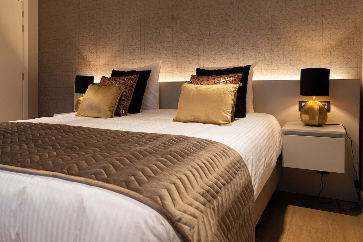 A neatly made bed with a patterned bedspread and multiple pillows, a bedside table with a lamp, and a wall with a geometric pattern.
