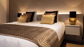 Een net opgemaakt bed met een geborduurd sprei en meerdere kussens, een nachtkastje met een lamp, en een muur met een geometrisch patroon.