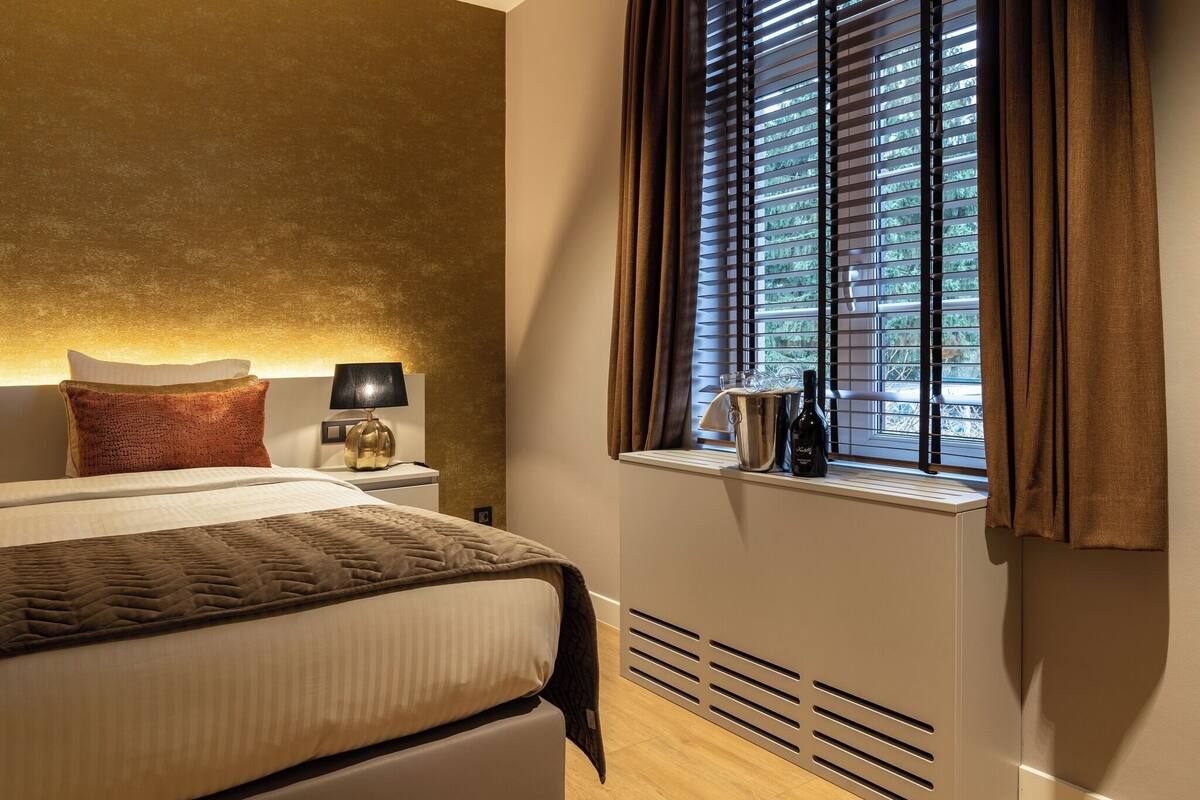 A hotel room with a bed, a nightstand, a lamp, a window with blinds, and a view of greenery outside.