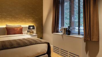 A hotel room with a bed, a nightstand, a lamp, a window with blinds, and a view of greenery outside.