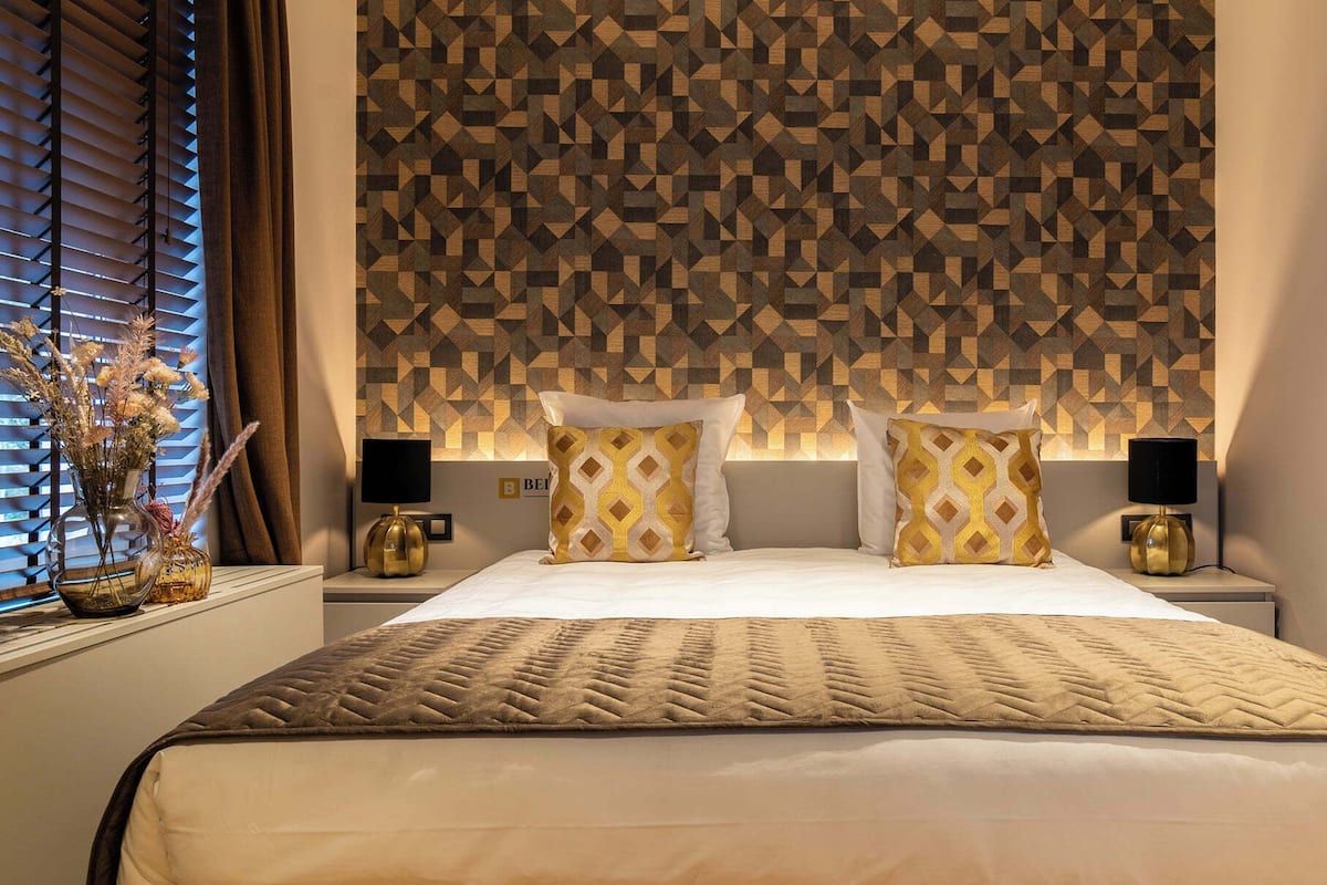 A bedroom with a geometric patterned headboard, two bedside tables with lamps, a bed with a patterned blanket, and a vase with flowers on the nightstand.