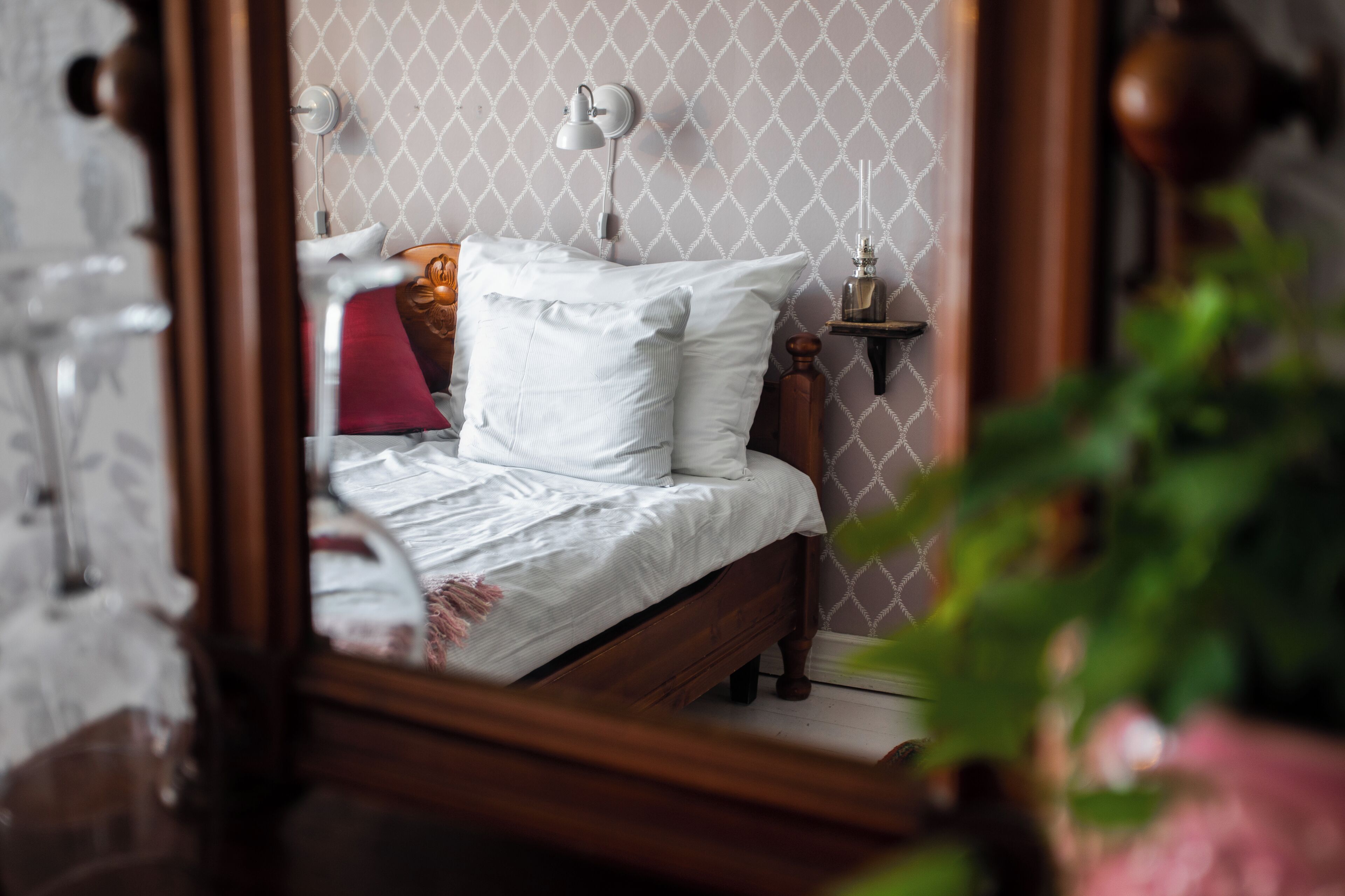 A bedroom with a wooden bed, white bedding, a bedside table with a lamp, and a wall-mounted mirror.