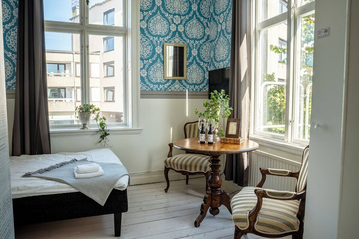 Une pièce comprenant un lit, une table ronde en bois avec des chaises, une plante en pot et une fenêtre donnant sur des immeubles.