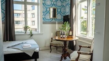 A room with a bed, a round wooden table with chairs, a potted plant, and a window with a view of buildings.