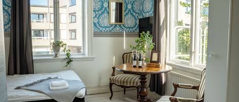 A room with a bed, a round wooden table with chairs, a potted plant, and a window with a view of buildings.