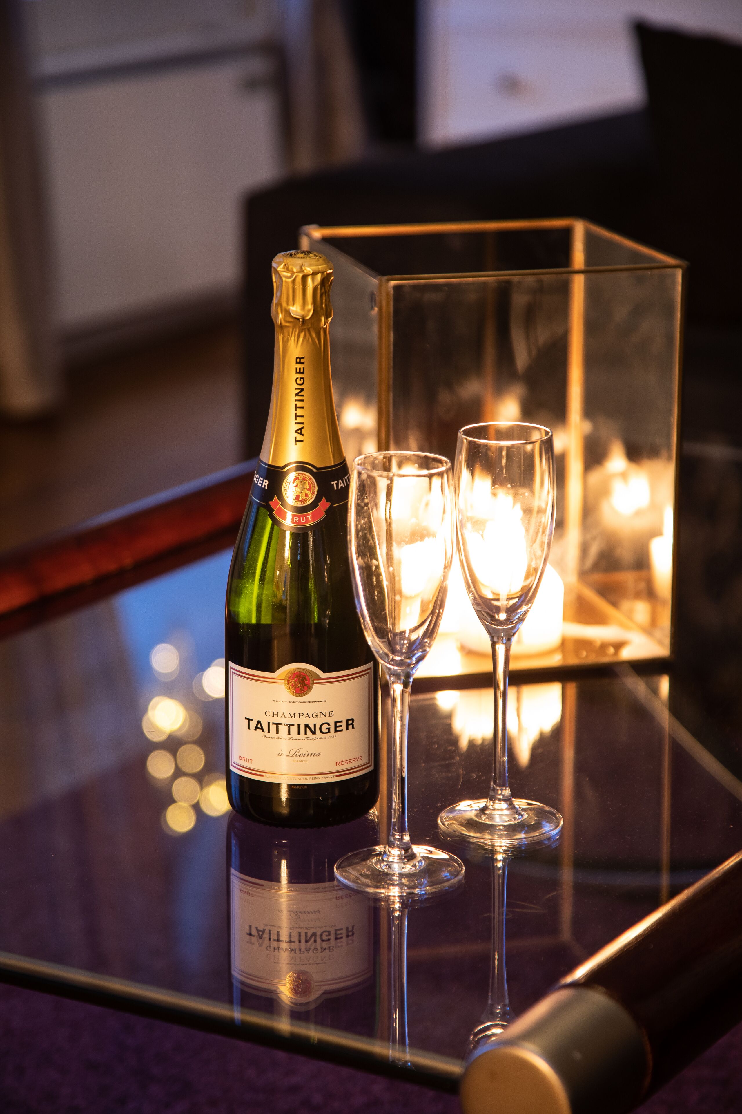 A bottle of Taittinger champagne and two glasses on a reflective surface with candles in the background.