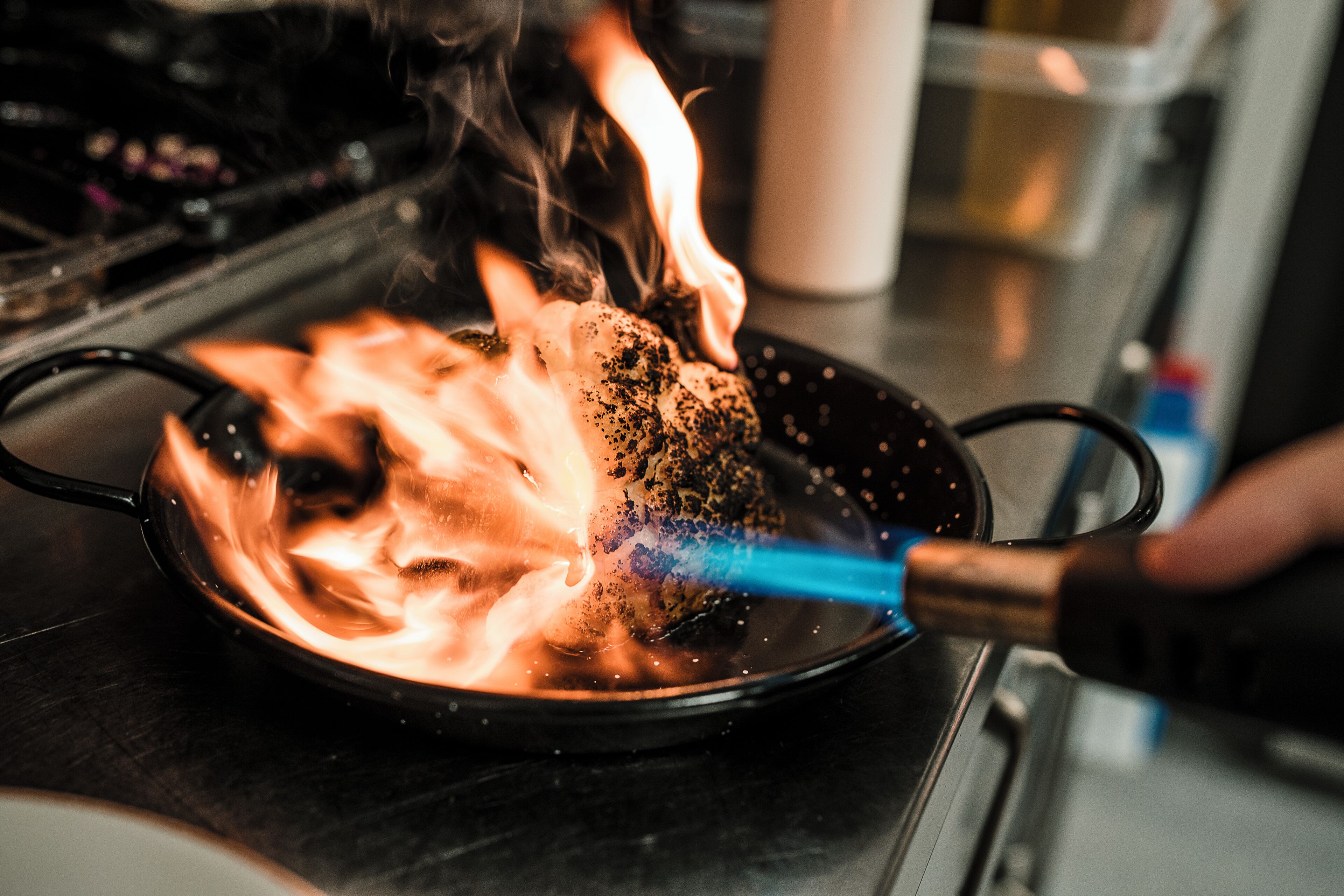 A close-up of a pan on a stove with flames and a blue tool.