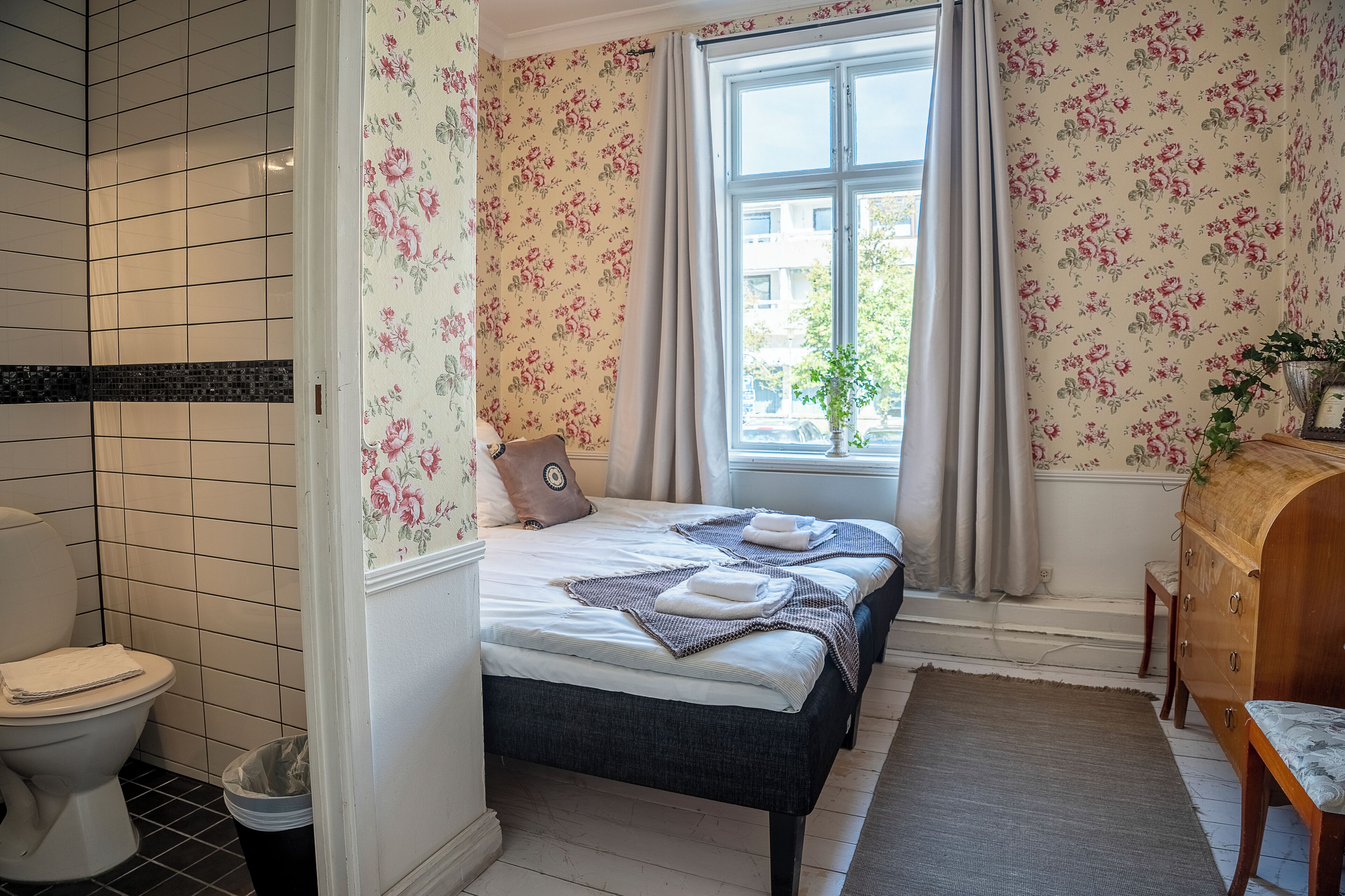 A bathroom with a toilet, a shower area, and a bed with floral wallpaper and a window.