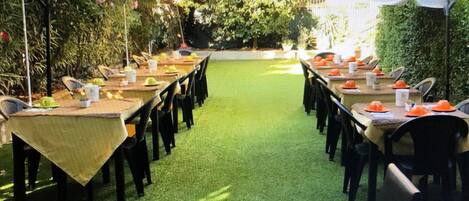 Un área de comedor al aire libre con mesas y sillas, cubierta por sombrillas y rodeada de vegetación.