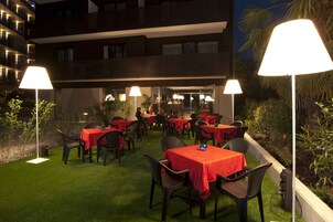 Un área de comedor al aire libre con mesas y sillas, rodeada de follaje e iluminada por lámparas.