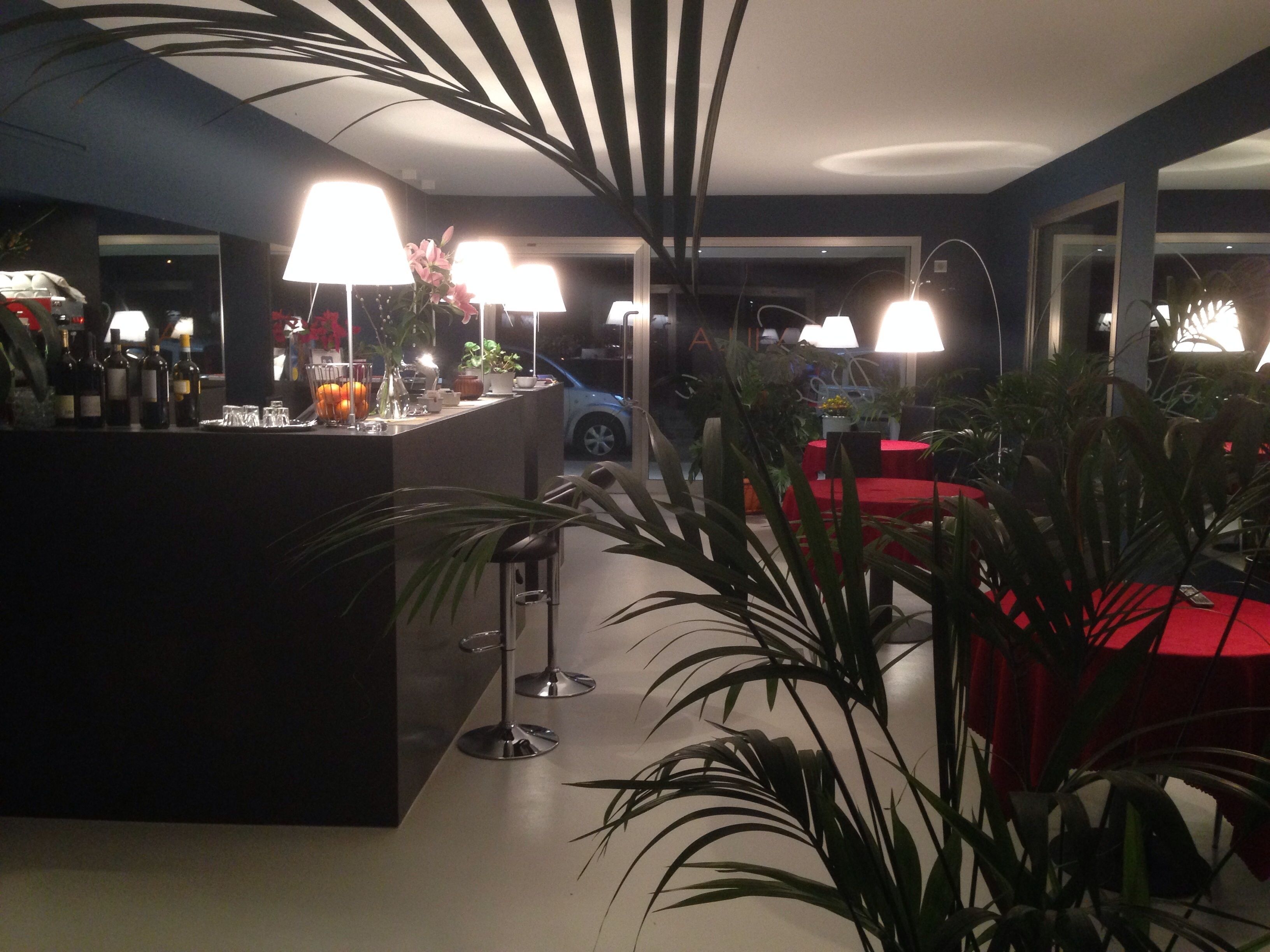 A modern bar area with a counter, wine bottles, glasses, and a potted plant. There are red tables and chairs in the background.