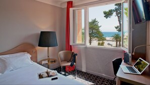 A hotel room with a bed, a desk with a laptop, a chair, a lamp, and a window with a view of the sea and trees. - Hôtel Beau Rivage (Argelès-sur-Mer)