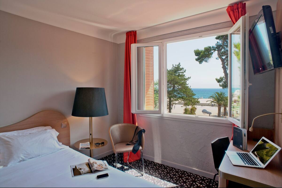 A hotel room with a bed, a desk with a laptop, a chair, a lamp, and a window with a view of the sea and trees.