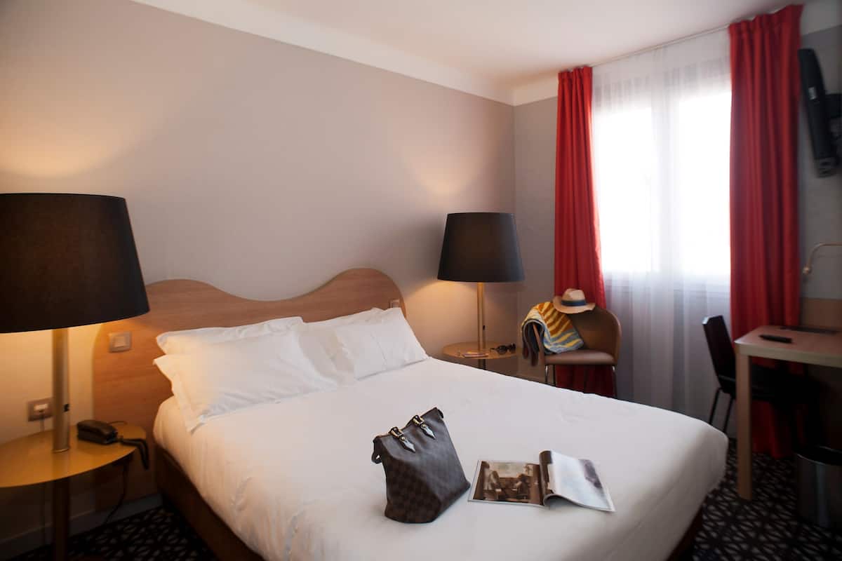 A hotel room with a bed, a desk, a chair, a lamp, and a handbag on the bed.