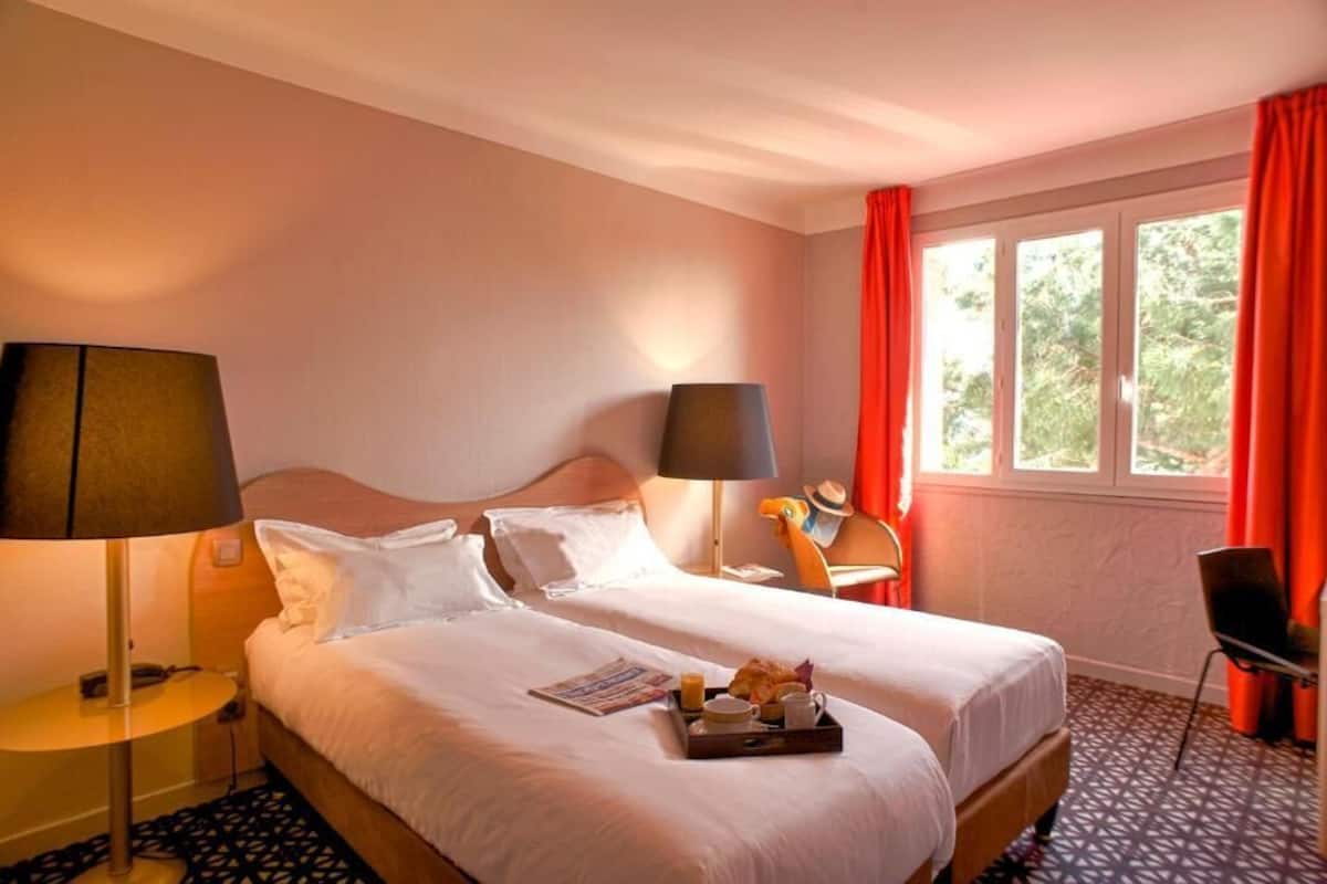 A hotel room with two beds, a window with red curtains, a small table with a lamp, and a chair.