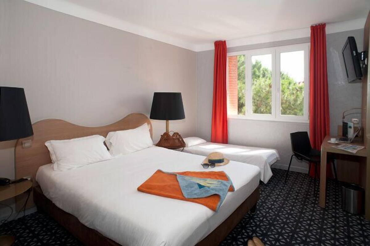 A hotel room with two beds, a desk, a chair, a TV, and a window with red curtains.