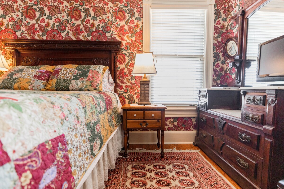 A bedroom with a floral-patterned bedspread, a wooden dresser, and a nightstand with a lamp.