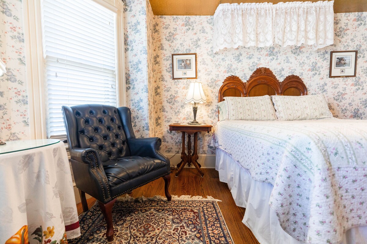 A bedroom with a floral wallpaper, a wooden bed with a white bedspread, a black leather chair, a small wooden table with a lamp, and a patterned rug.