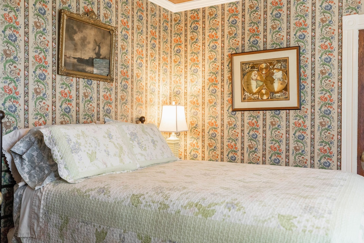 A bedroom with a floral wallpaper, a bed with a quilt, two framed pictures, and a bedside lamp.