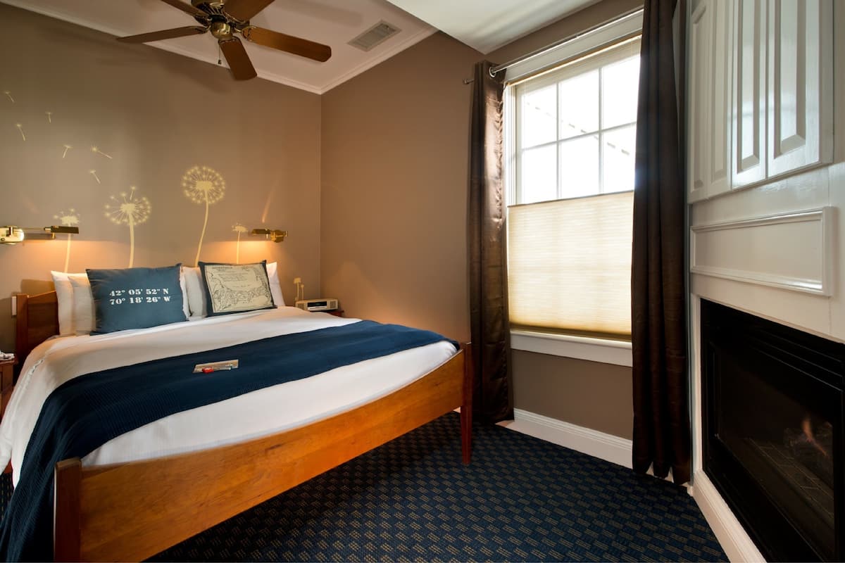 A hotel room with a wooden bed, a fireplace, a large window with curtains, and wall-mounted lamps.