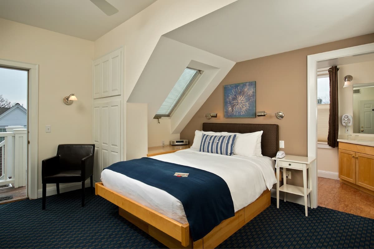 A bedroom with a bed, a chair, a nightstand, a painting, and a bathroom visible through an open door.