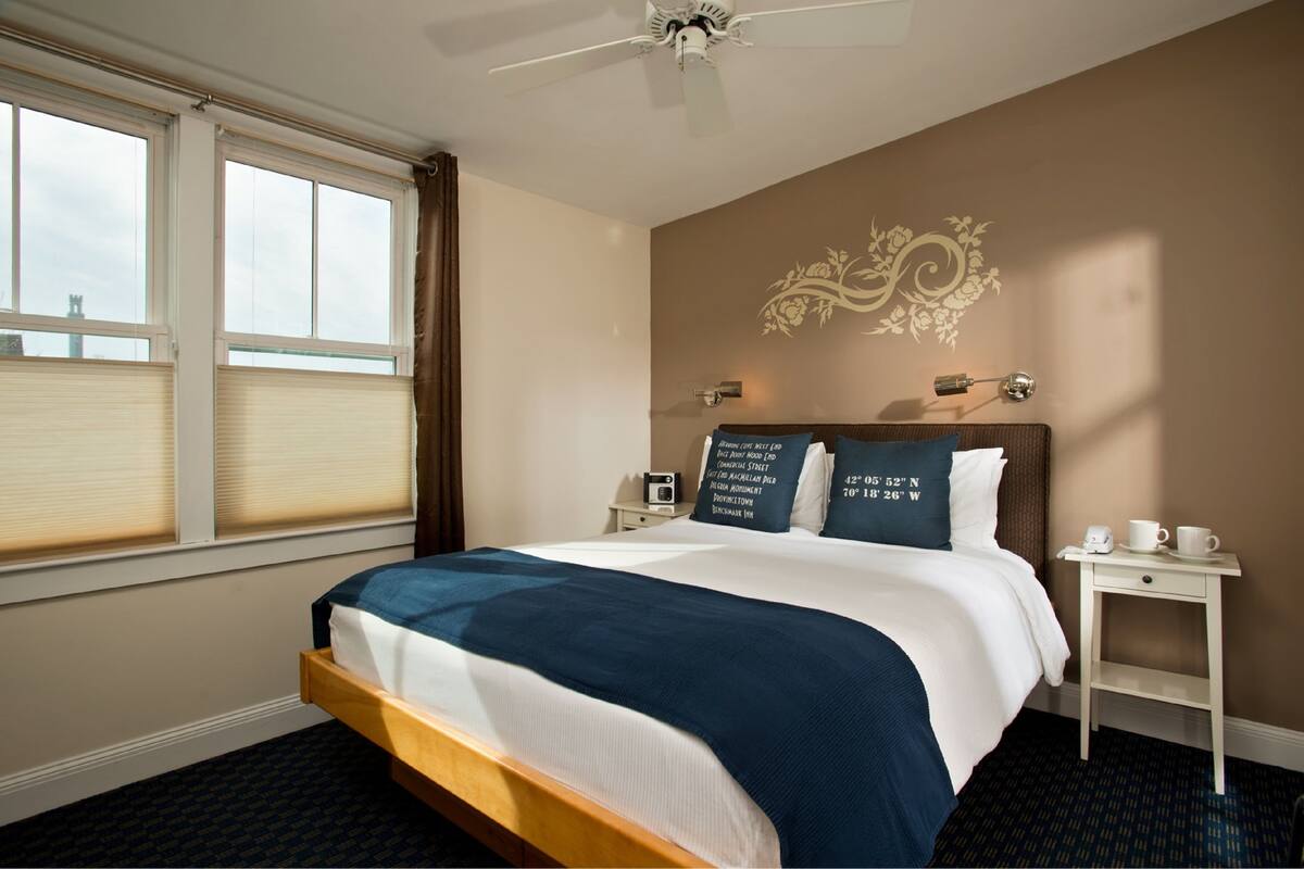 A hotel room with a bed, a nightstand, a wall clock, and a window with blinds.