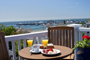 Una mesa de madera con alimentos y bebidas para el desayuno en un balcón con vistas a un puerto deportivo con barcos y un muelle.