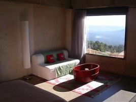 Una habitación con cama, un pequeño sofá con cojines, un cubo rojo y una ventana con vistas a montañas.