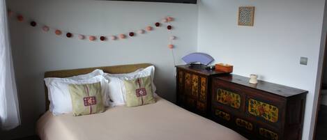Una cama bien hecha con una colcha color beige y almohadones decorativos, un aparador de madera con motivos florales, y una guirnalda de bolas rosas y melocotón en la pared.