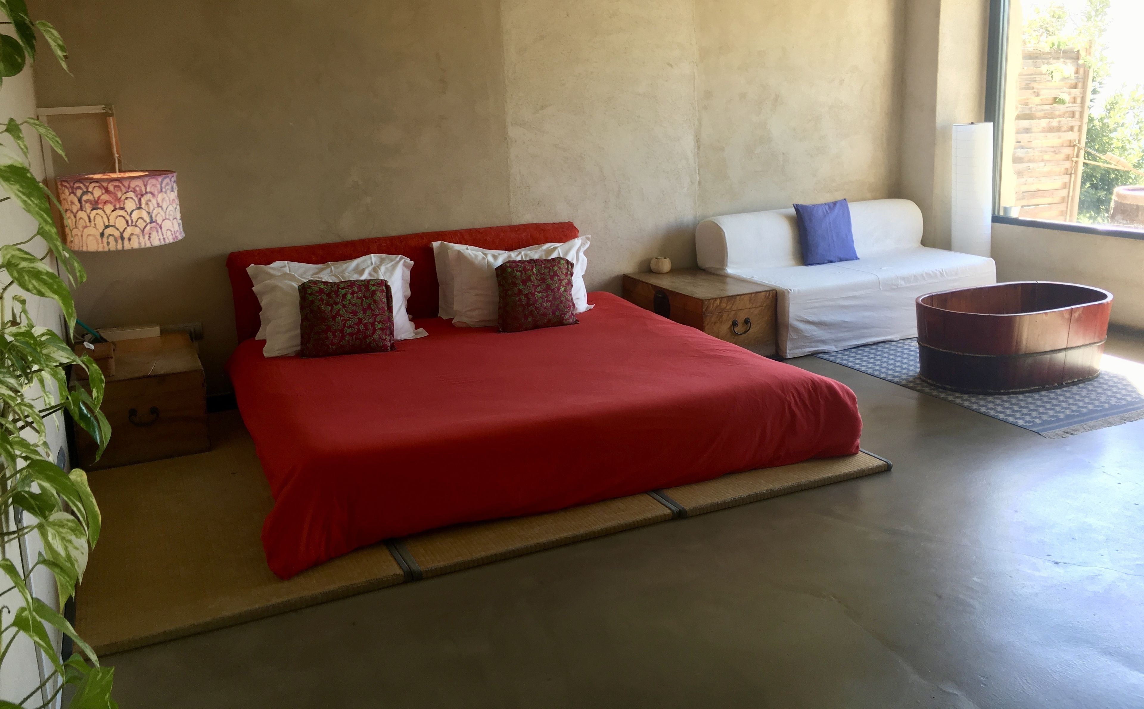 A bedroom with a red bed, white pillows, a wooden nightstand, a white sofa, and a copper bucket.
