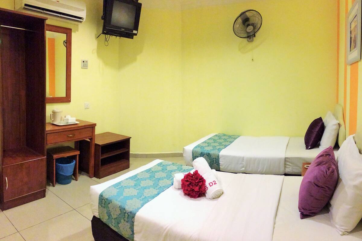 A hotel room with two beds, a desk, a chair, an air conditioner, a television, and a wall fan.